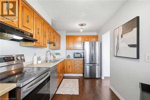 Kitchen featuring wood cabinetry, stainless steel appliances, a double basin sink, and wood-finish flooring - 375 King Street N Unit# 708, Waterloo, ON - Indoor Photo Showing Kitchen