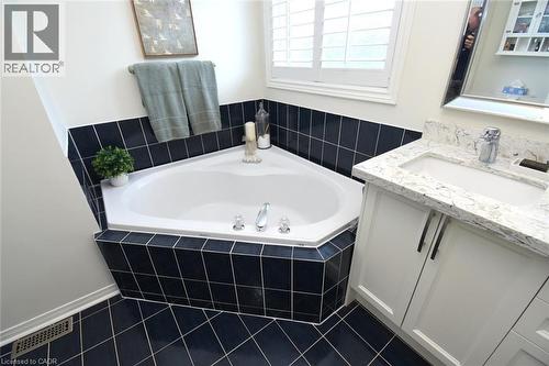 Corner soaking tub with dark tile surround, white shaker-style vanity with stone-finish countertop, under-mount sink, and window with plantation shutters - 470 Amanda Crescent, Burlington, ON - Indoor Photo Showing Bathroom