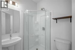 White pedestal sink with a chrome faucet, mirrored vanity, glass-enclosed shower, white toilet, and a wall-mounted wood-finish shelf - 