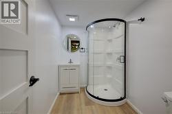 Curved glass shower enclosure with black trim, vanity with integrated sink, wood-finish flooring, and a circular mirror - 