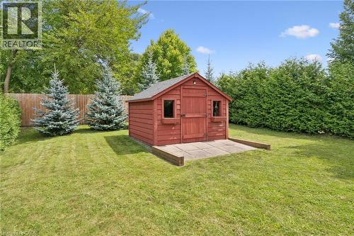 Red wood-sided storage shed on a concrete pad, surrounded by a grassy lawn, mature evergreen trees, and a wood fence - 49 East Street, Cambridge, ON - Outdoor