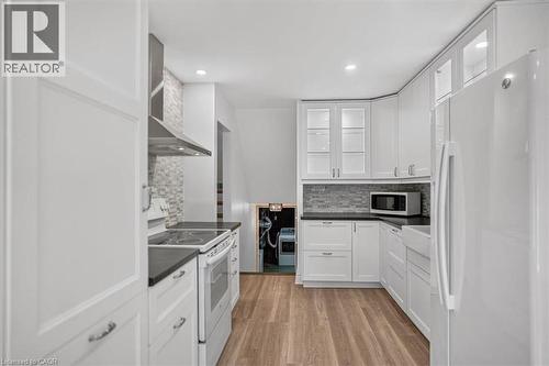 White cabinetry with glass-front uppers, dark countertops, a tile backsplash, stainless steel range hood, and wood-finish flooring - 27 Gretna Court, Hamilton, ON - Indoor Photo Showing Kitchen