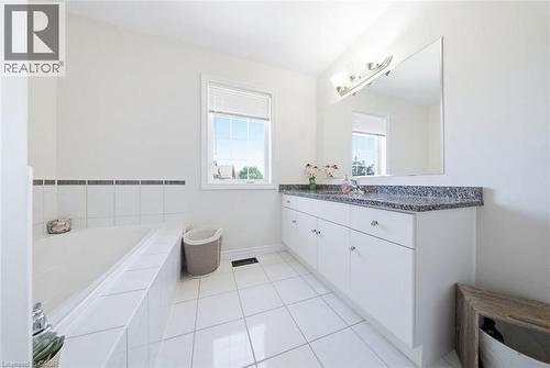 White tile flooring, a built-in bathtub with a tiled surround, a window with blinds, a vanity with a dark speckled countertop, and a wall-mounted mirror - 65 Amsterdam Crescent, Guelph, ON - Indoor Photo Showing Bathroom