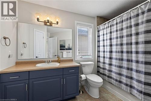 Vanity with a built-in sink and dark blue cabinetry, bronze-finish wall-mounted light fixture, large wall mirror, toilet, and tile flooring - 783 Frontenac Crescent, Woodstock, ON - Indoor Photo Showing Bathroom