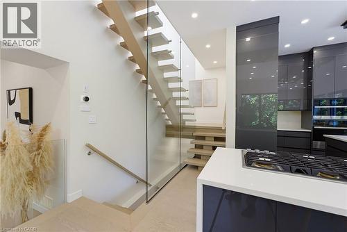 Open-tread staircase with glass railing, light wood-finish flooring, recessed lighting, and a modern kitchen featuring dark high-gloss cabinetry and a built-in gas cooktop - 282 Robina Road, Ancaster, ON - Indoor Photo Showing Other Room
