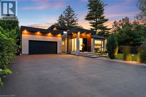 Contemporary architectural design featuring a flat roofline, integrated exterior lighting, a dark-paneled garage door, a paved driveway, and mature perimeter landscaping - 282 Robina Road, Ancaster, ON - Outdoor