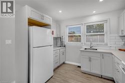 Modern kitchen featuring light gray cabinetry, light countertops, wood-finish flooring, white subway tile backsplash, and a double window with blinds - 