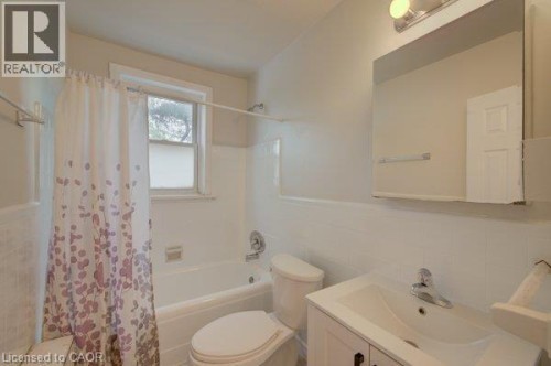 White ceramic tile walls, built-in bathtub with shower fixture, single-pane window, white vanity with integrated sink, and wall-mounted mirror - 141 Chapel Street, Kitchener, ON 