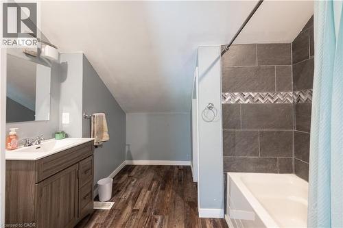 Wood-finish flooring, single vanity with integrated sink, modern wall sconce, tiled shower surround with decorative chevron accent, and white bathtub - 146 Benson Street, Ingersoll, ON 