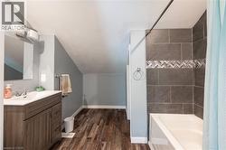 Wood-finish flooring, single vanity with integrated sink, modern wall sconce, tiled shower surround with decorative chevron accent, and white bathtub - 