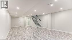 Spacious room featuring wood-finish flooring, recessed lighting, a white paneled door, and a staircase with a black handrail and glass balustrade - 