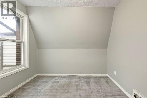 Slanted ceiling design, light gray wall finish, white window trim, gray carpet flooring, and white baseboards - 23 Frederick Avenue, Hamilton, ON 