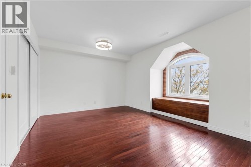 Room featuring rich wood-finish flooring, a large arched window with a built-in wood bench, white painted walls, and a contemporary flush-mount ceiling light - 1389 Lakeshore Road, Burlington, ON 