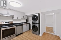 Modern kitchen and laundry space featuring stainless steel appliances, white cabinetry with hardware accents, wood-finish flooring, a dual-stack washer and dryer unit with black finish, and recessed c - 