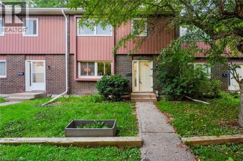 Welcoming townhome entry featuring a brick and metal-panel exterior, mature tree-lined landscaping, and a manicured front lawn with established plantings - 121 University Avenue E Unit# 102, Waterloo, ON 