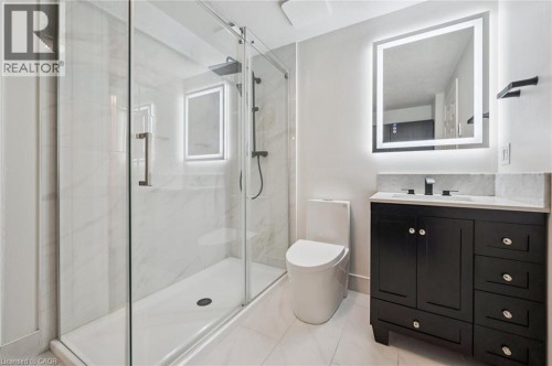 Shower with clear glass enclosure and rainfall showerhead, black vanity with an integrated sink, illuminated mirror, and a white toilet - 426 Hidden Creek Drive, Kitchener, ON 
