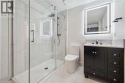 Shower with clear glass enclosure and rainfall showerhead, black vanity with an integrated sink, illuminated mirror, and a white toilet - 