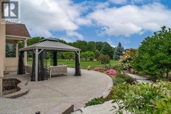 Expansive patio featuring an exposed aggregate finish, a metal-roofed gazebo with privacy screens, a built-in stone fire pit, and landscaped garden beds with mature plantings - 