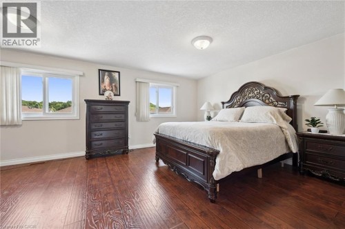 Spacious primary bedroom featuring hardwood flooring throughout, ornate dark wood bed frame with upholstered headboard, dual nightstands with attached lighting, and multiple windows with natural light - 86 Bridlewreath Street, Kitchener, ON 