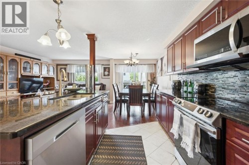 Kitchen featuring dark wood cabinetry, stainless steel appliances, a tile backsplash, and a central island with a built-in sink and stone countertop - 86 Bridlewreath Street, Kitchener, ON 