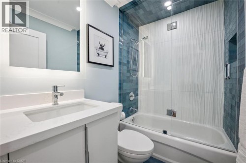 Contemporary vanity with integrated sink and chrome faucet, backlit mirror, and a shower-tub combination featuring textured white tile and blue mosaic tile accents - 1261 Stephenson Drive, Burlington, ON 