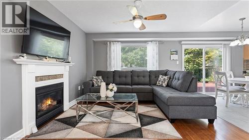 Fireplace with a classic white mantel, wood-finish flooring, ceiling fan, sliding glass door, and a kitchen visible in the background - 124 Osborn Avenue, Brantford, ON 
