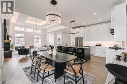 Open-concept living area featuring light wood-finish flooring, a contemporary kitchen with white cabinetry and a black island, a dining space with a unique chandelier, and a living room with large win - 