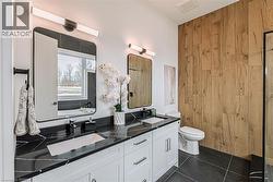 Dual vanity with black countertops and integrated sinks, white cabinetry, wood-finish accent wall, dark floor tiling, and a separate toilet - 