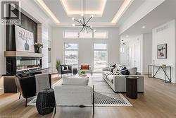 Tray ceiling with recessed lighting and a contemporary chandelier, linear fireplace with a black surround and wood mantel, large windows, wood-finish flooring, and white walls - 