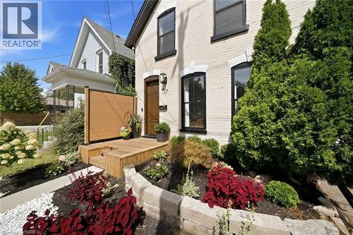 White brick exterior with black window trim, wooden entry door, elevated wooden porch, and landscaped garden beds - 57 Terrace Hill Street, Brantford, ON 