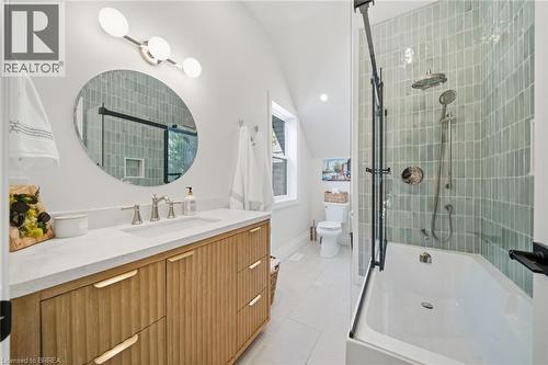 White porcelain bathtub with glass enclosure, vertical tile shower surround, wood-finish vanity with integrated sink, and a circular mirror - 57 Terrace Hill Street, Brantford, ON 