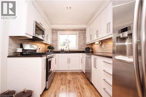 Galley kitchen featuring wood-finish flooring, white cabinetry with stainless steel hardware, dark countertops, and a tiled backsplash - 380 Grangewood Drive, Waterloo, ON 