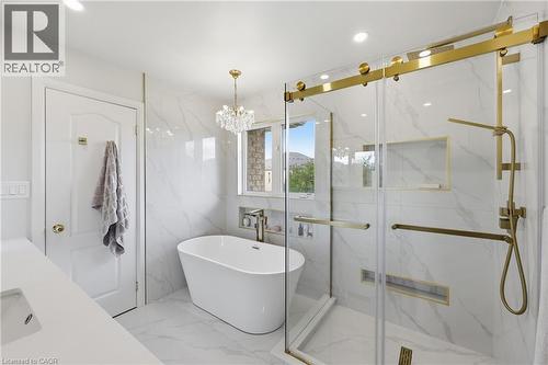 Freestanding soaking tub with a crystal chandelier, glass-enclosed shower with gold-toned hardware, white marble-look wall and floor tiling, and a recessed window - 738 Fourth Line, Milton, ON - Indoor Photo Showing Bathroom