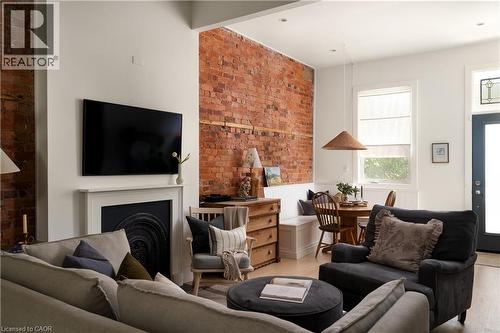 Exposed brick accent wall, built-in banquette seating, a white painted fireplace with a decorative insert, and light wood-finish flooring - 29 Steven Street, Hamilton, ON 