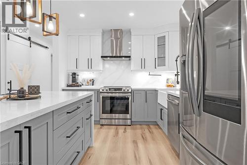 Contemporary kitchen featuring a large central island with light gray cabinetry, stainless steel appliances, wood-finish flooring, and a white farmhouse sink - 4236 Longmoor Drive, Burlington, ON 
