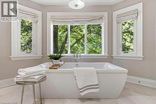 Freestanding soaking tub with polished chrome fixtures, surrounded by three large windows, featuring neutral-toned wall paint and light-toned flooring - 4038 Lakeshore Road, Burlington, ON 