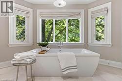 Freestanding soaking tub with polished chrome fixtures, surrounded by three large windows, featuring neutral-toned wall paint and light-toned flooring - 