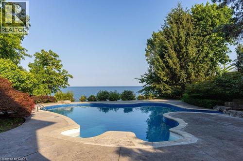 Freeform swimming pool with a stamped concrete surround, mature perimeter landscaping, and a substantial body of water in the background - 4038 Lakeshore Road, Burlington, ON 