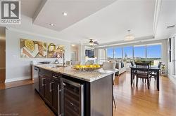 Open-concept living area with wood-finish flooring, a kitchen island featuring a beverage cooler and granite countertop, and a tray ceiling with recessed lighting - 