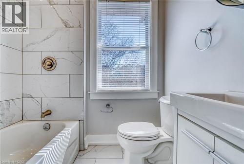 Bathroom featuring a white bathtub, marble-patterned tile shower surround, matching floor tile, built-in vanity with integrated sink, and white window blinds - 23 Gibson Avenue, Hamilton, ON 