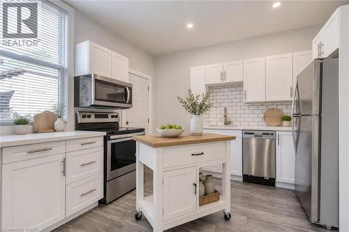 Kitchen featuring wood-finish flooring, white cabinetry with modern hardware, stainless steel appliances, a subway tile backsplash, and a movable island with a wood countertop - 23 Gibson Avenue, Hamilton, ON 