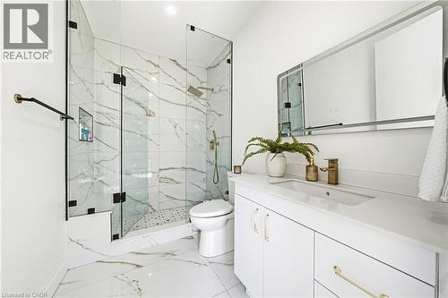 Floor-to-ceiling marbled tile, glass-enclosed shower with gold-tone fixtures, white vanity with integrated sink, and a framed mirror - 4511 Guelph Line, Burlington, ON 