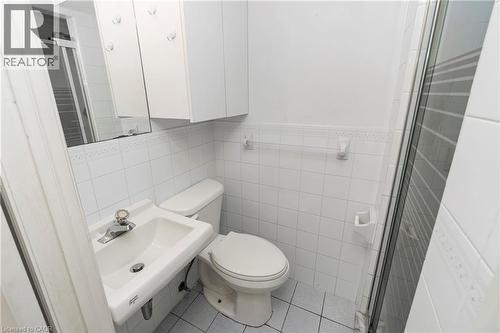 White ceramic tile walls, white wall-mounted sink, white toilet, wall-mounted storage cabinet, and a glass-enclosed shower - 36 Buckingham Drive, Hamilton, ON 
