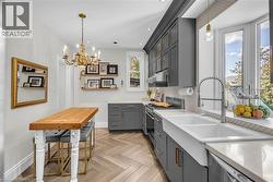 Herringbone wood-finish flooring, grey cabinetry with gold-tone hardware, white farmhouse sink, white countertops, and a large bay window - 