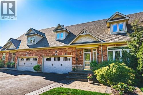 Brick exterior with multiple dormer windows, attached two-car garage, paved driveway, and stone-paver walkway - 31 Book Road Unit# 44, Grimsby, ON 