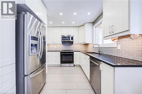 Modern kitchen featuring white cabinetry, stainless steel appliances, dark countertops, a subway tile backsplash, and light-toned tile flooring - 2037 Mountain Grove Avenue, Burlington, ON 