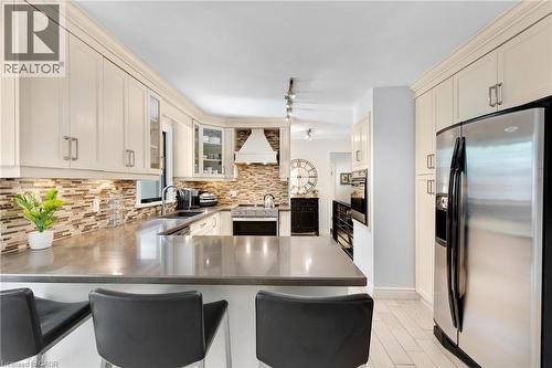 Kitchen featuring cream-colored cabinetry, stainless steel appliances, a mosaic tile backsplash, a range hood, and a built-in kitchen island with seating - 7 Shadywood Lane, Cambridge, ON 