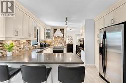 Kitchen featuring cream-colored cabinetry, stainless steel appliances, a mosaic tile backsplash, a range hood, and a built-in kitchen island with seating - 