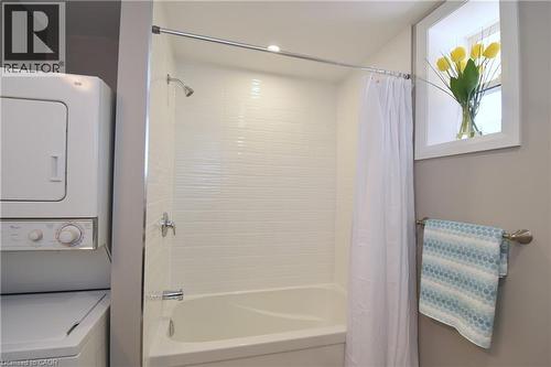 White stacked washer and dryer, white tiled shower surround, white bathtub, chrome shower fixture, and a window with white trim - 76 Burlington Street E, Hamilton, ON 