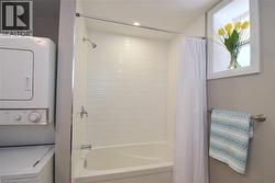 White stacked washer and dryer, white tiled shower surround, white bathtub, chrome shower fixture, and a window with white trim - 